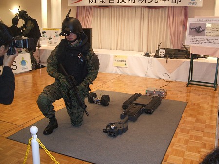 しゃがんだ姿勢で構える「先進個人装備」を装備する隊員。右側に転がっているのは「携帯型ロボット」(後述)